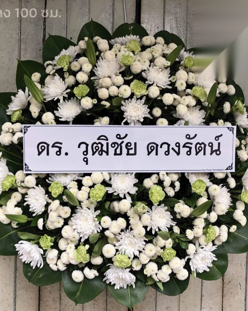 พวงหรีดดอกไม้สดโทนขาวเขียวทรงกลม ป้ายชื่อผู้ส่งสีขาวข้อความสีดำ ตั้งพิงผนังไม้ระแนง